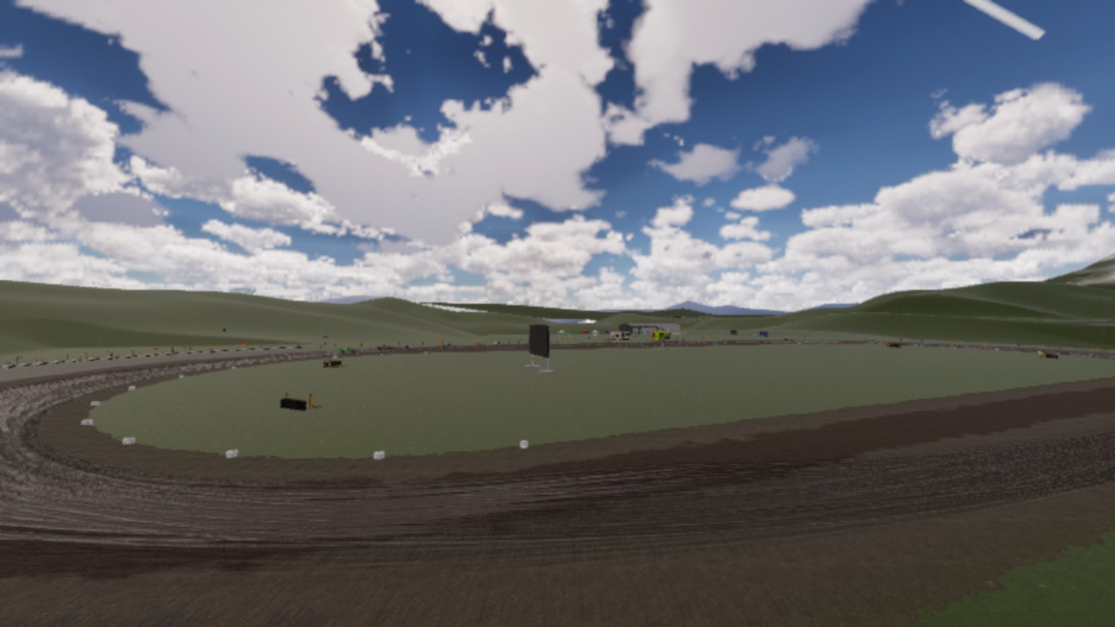 A wide view across an oval dirt racing track in a grassy valley under a bright sky full of white cloud. Small white markers line the inside of the track, a tall dark screen stands on a frame in the infield, and buildings and tents sit far off on the other side.