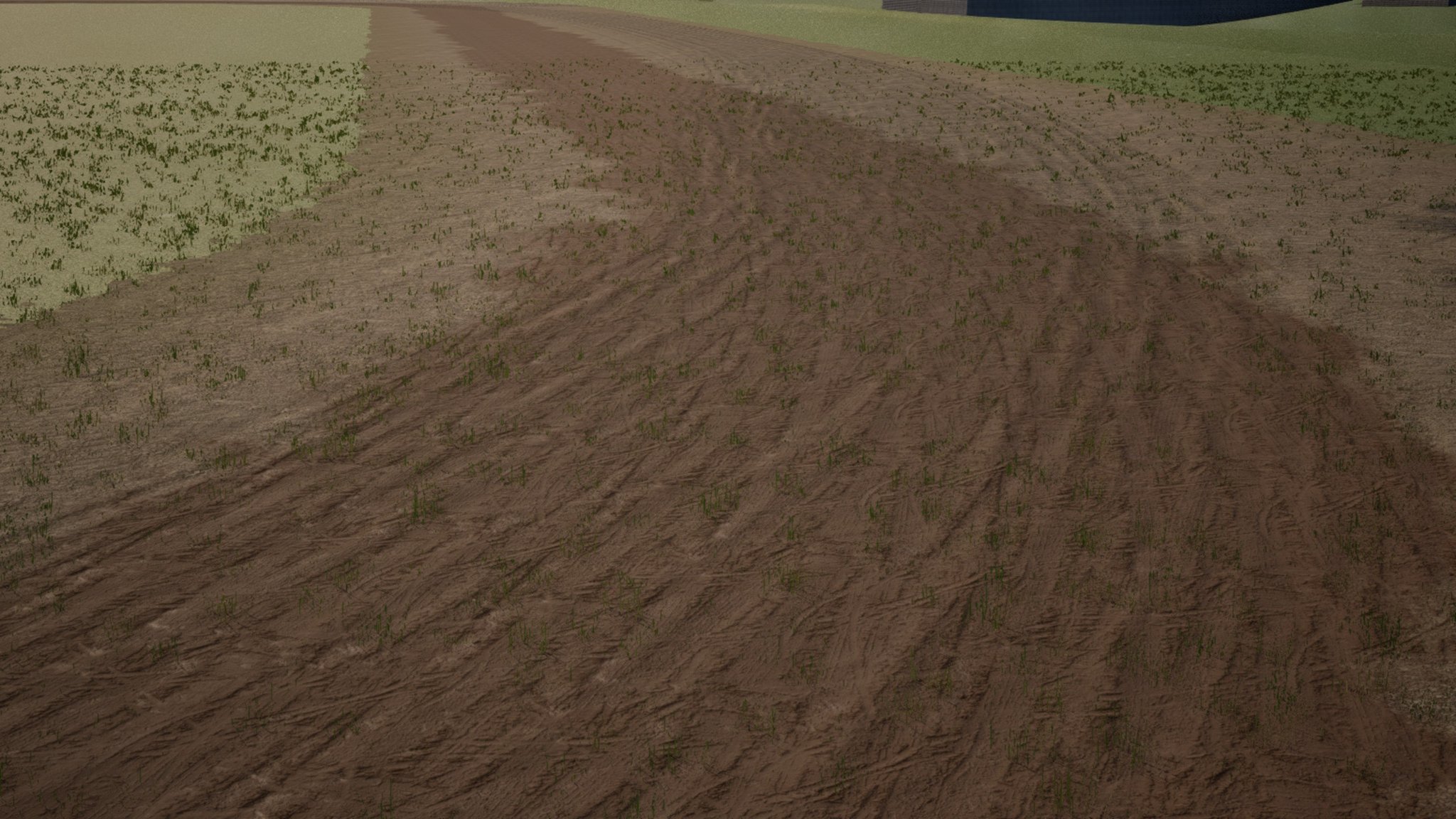 A low view along a brown mud racing track cut into a grass field. A darker, dished band sweeps through the bend, covered in overlapping tyre grooves, with tufts of grass poking through the mud.
