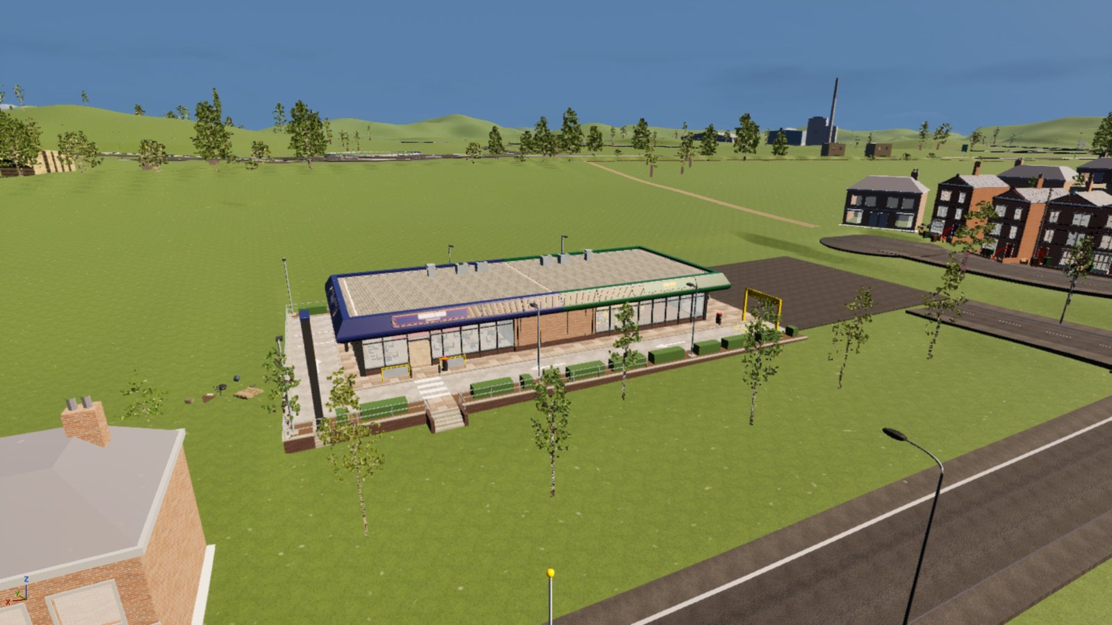 An aerial view of a single long flat-roofed drive-thru building on open green ground, one half with a navy fascia and the other with a green one, a paved frontage with planters, steps and a zebra-striped crossing, a yellow height gantry at the end of the drive lane and a tarmac car park beside it. Red-brick houses and a road are to the right, and hills with trees and a tall chimney fill the horizon.