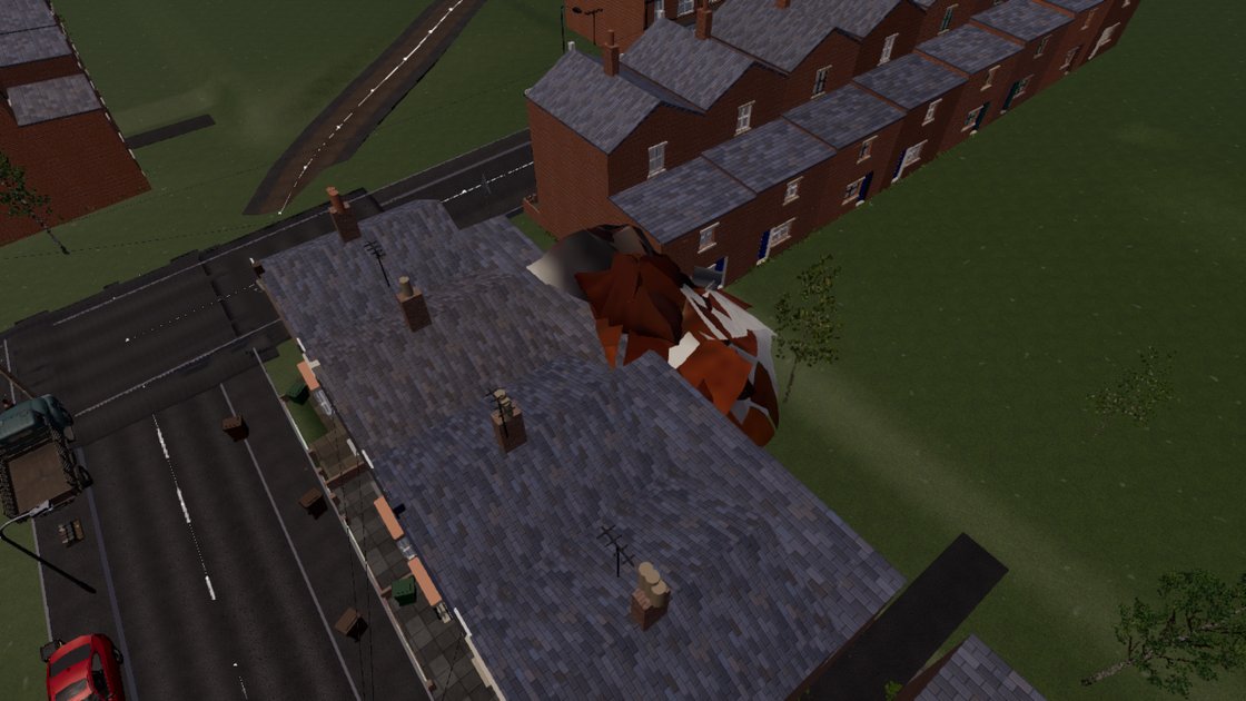 An aerial view over a row of terraced roofs, a road and green fields. Above the roofs floats a huge shattered shape of brown, white and red shards, bigger than a house, which is a single hen mesh blown apart by its animation.
