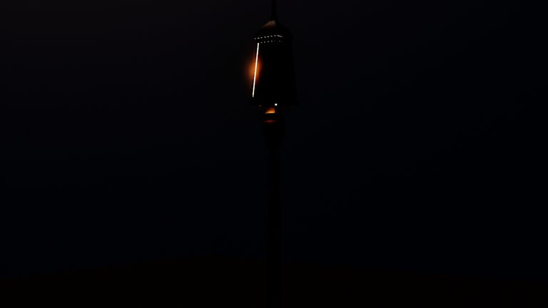 A tall black cast-iron lamp post at night on a tiled floor. The lantern at the top reads as a solid dark box with a thin bright line of orange light escaping down one corner seam and along its base, while a wide pool of warm light falls on the floor in the foreground.