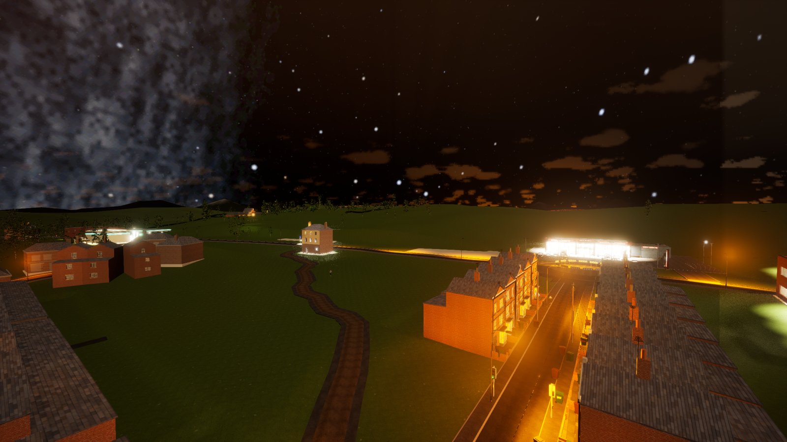 The town from above at night: a terraced street lit orange by its lamps, a lit supermarket, green fields and a winding path. Small clouds glow orange-brown from below, and on the left a large blotchy grey patch of stars fills the sky.