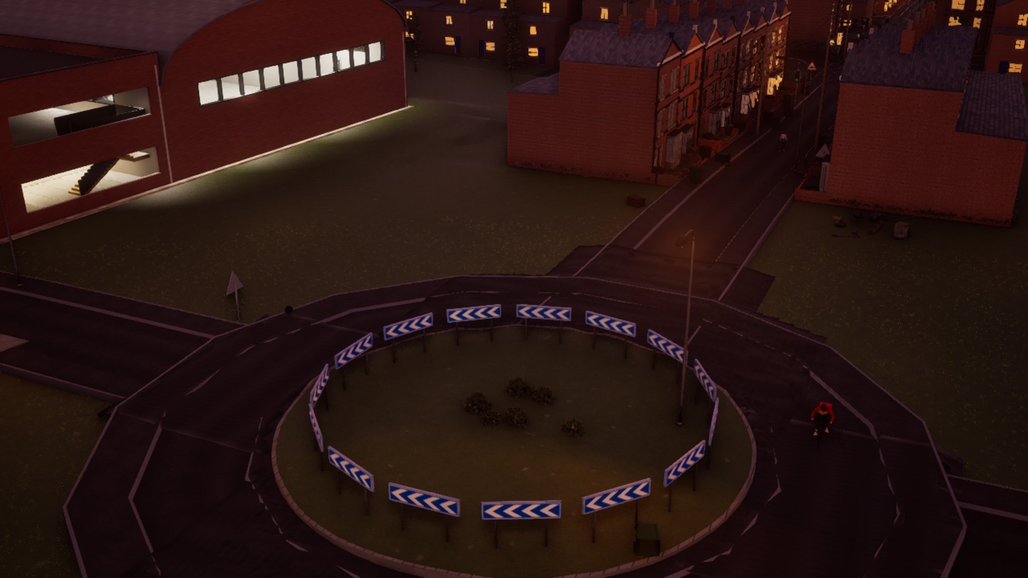 A roundabout at night from above: a ring of blue and white chevron signs around a grassed centre with a few shrubs, the circulating road and its arms around it, terraced houses with lit windows behind.