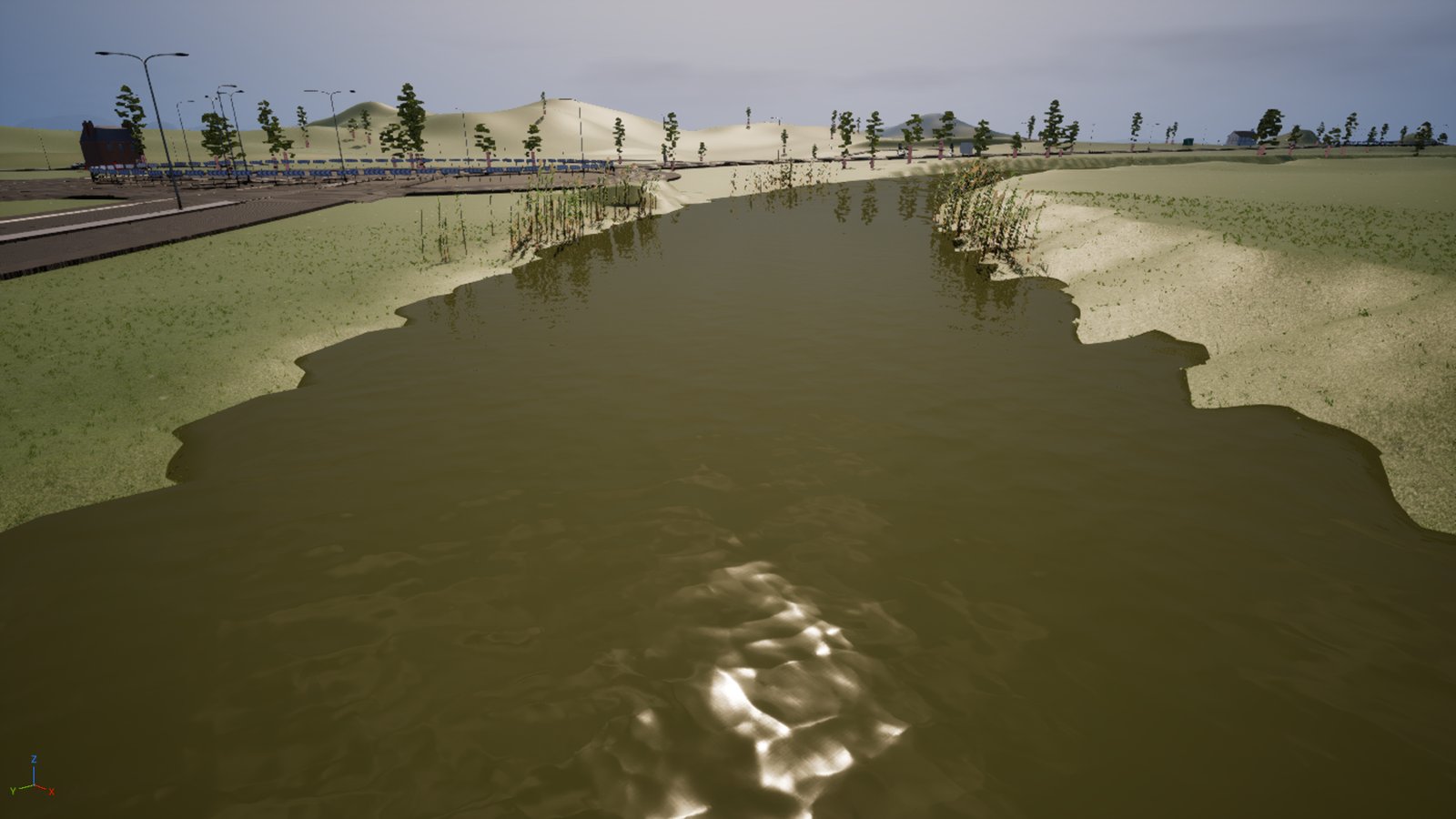 A river seen from above the bank in daylight, the water a murky olive-brown with a sun glare on the surface, reeds standing along both margins, a road and railings on the left and low hills behind.