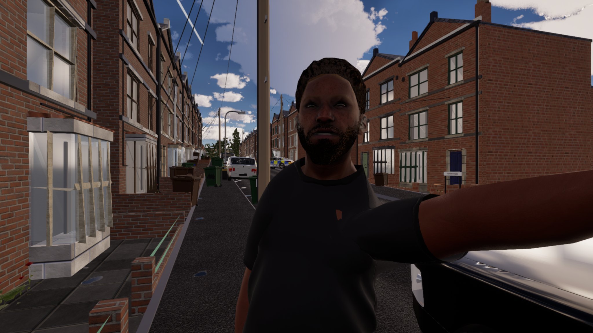 A bearded man in a black T-shirt stands close in front of the viewer on a terraced street in daylight, his left arm reaching out of frame towards the viewer's shoulder, a parked car beside him.
