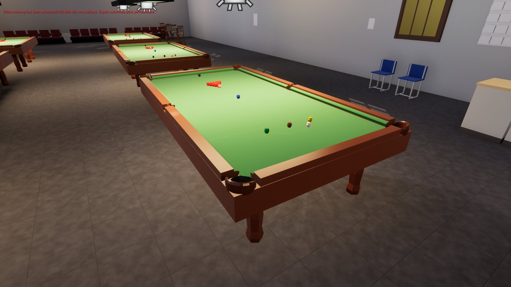 A lit snooker hall with three full-size tables in a row on a grey tiled floor. The nearest table is part way through a frame - the reds are still roughly bunched but spread at the edges, the blue sits on its spot, and the brown, green and white are down near the baulk end. Blue stacking chairs and a bar counter stand against the right-hand wall, rows of red seating at the far end. A red engine warning across the top left reads that video memory has been exhausted.